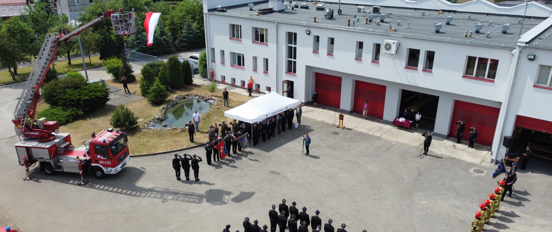 Zdjęcie przedstawia widok uroczystości, która ma miejsce na placu wewnętrznym komendy Powiatowej PSP w Krośnie Odrzańskim z lotu ptaka. Na zdjęciu widoczne są pojazdy oraz pododdziały oddające honory podczas podniesienia flagi państwowej.