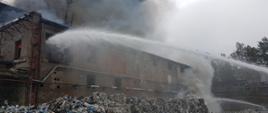 Zdjęcie przedstawia palący się budynek hala magazynowa, przed budynkiem znajduje się sterta nie spalonych śmieci. Na budynek podawane są dwa prądy gaśnicze jeden na palące się śmieci drugi do środka budynku przez okno na 2 kondygnacji