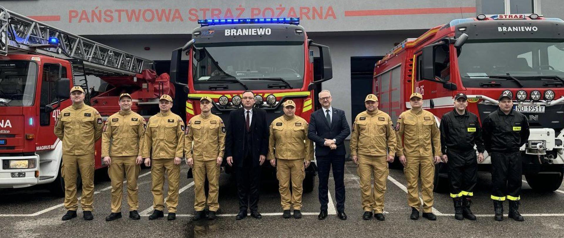 Na zdjęciu obok siebie stoją Wojewoda Warmińsko mazurski, burmistrz Braniewa ze strażakami. W tle samochody strażackie i remiza JRG Braniewo