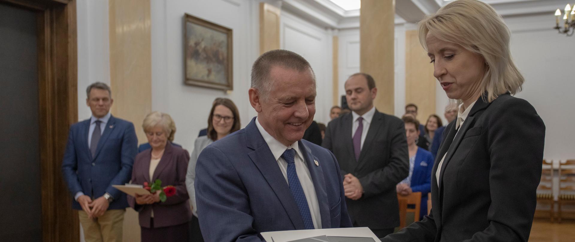 Minister Teresa Czerwińska wręcza medal szefowi KAS Marianowi Banasiowi, w tle pozostali nagrodzeni w sali w MF