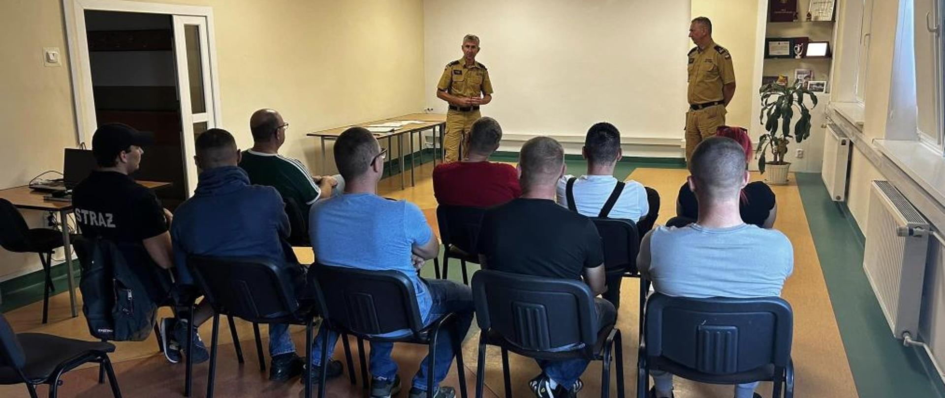 Widok na sale szkoleniową, świetlice. Na środku sali siedząca na krzesłach grupa ludzi. Przed grupką stoi dwóch strażaków ubranych w piaskowe mundury. Tłumaczą coś zgromadzonej grupie. Osoby widoczne są od tyłu a prowadzące od przodu.