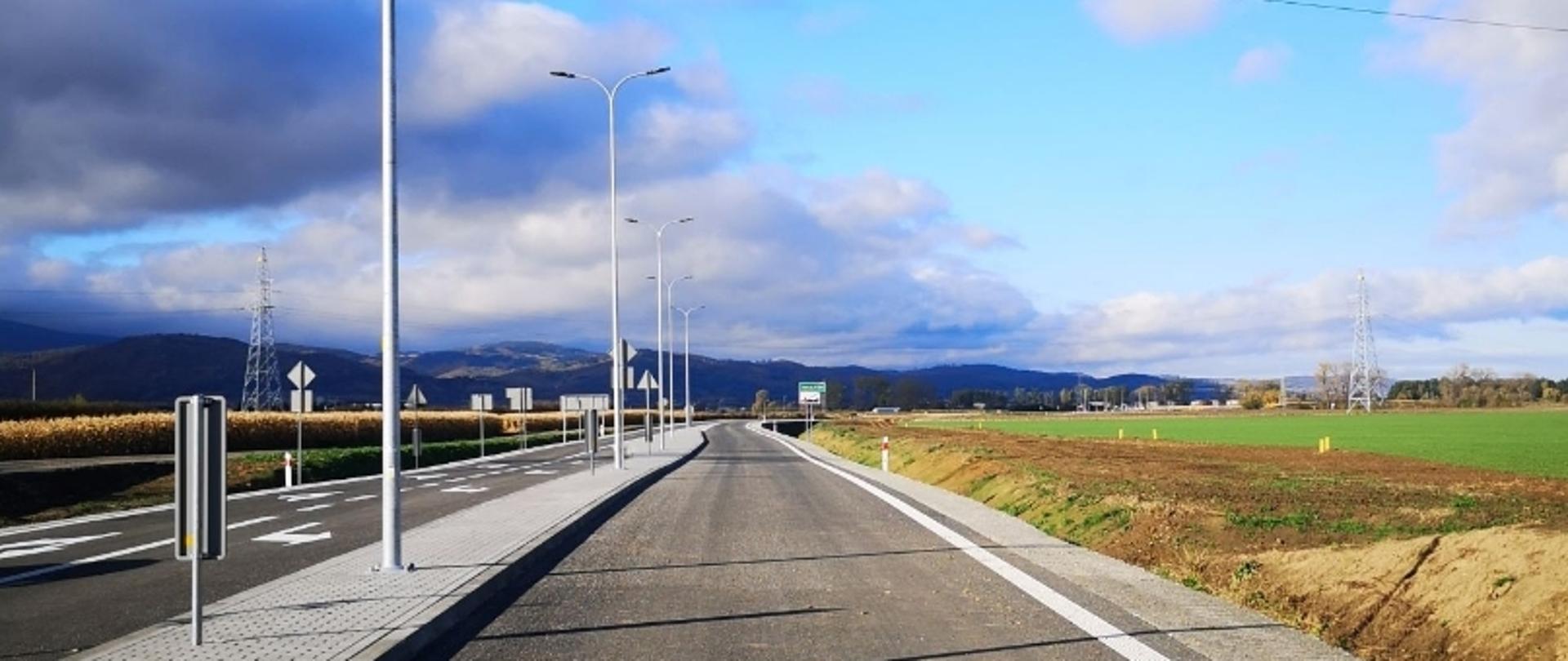 Nowa droga z latarniami i znakami, otoczona polami, z górami w tle.