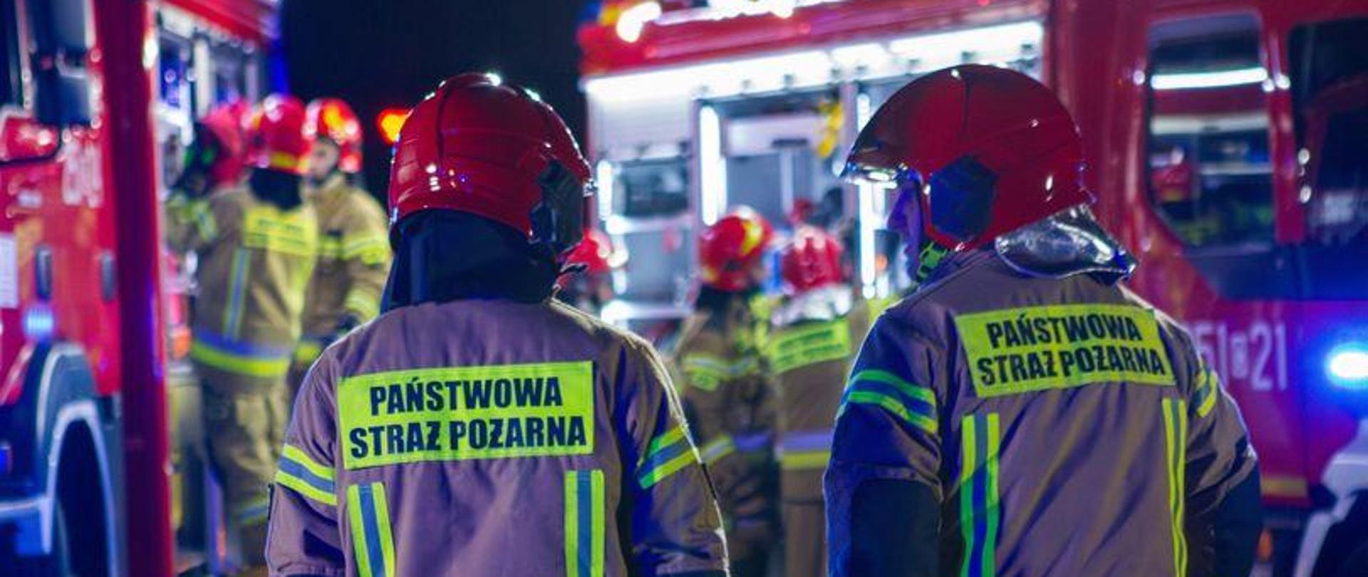 Strażacy podczas działań ratowniczo-gaśniczych. W tle widoczne pojazdy pożarnicze i strażacy.