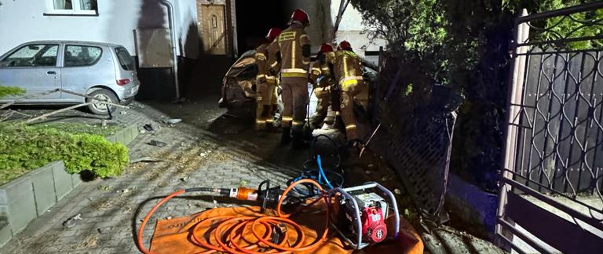 Na zdjęciu widać pole sprzętowe strażaków oraz torbę medyczną. W tle strażacy wykonują dostęp do poszkodowanego poprzez drzwi pasażera.