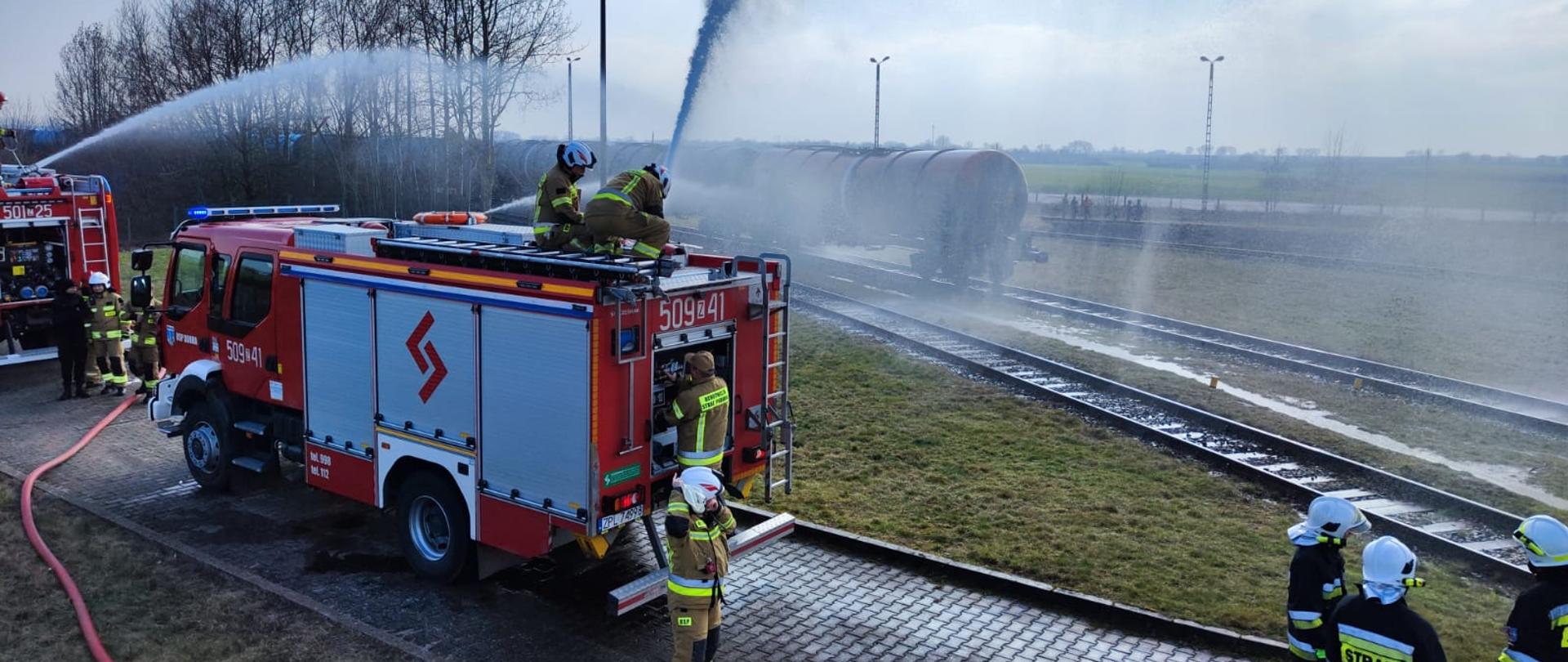 Na zdjęciu po lewej stronie widoczne z tyłu jeden za drugim dwa czerwone samochody pożarnicze ustawione na utwardzonej nawierzchni z kostki polbruku. Z ich dachów podawane silne strumienie wody na ostatni wagon stojącego na torze składu cystern kolejowych. Na dachu, przy pojazdach i na drodze stojące grupki strażaków ubranych w czarne i w piaskowe mundury i białe hełmy. W tle bezlistne drzewa, jasne niebo z niewielkimi obłokami.