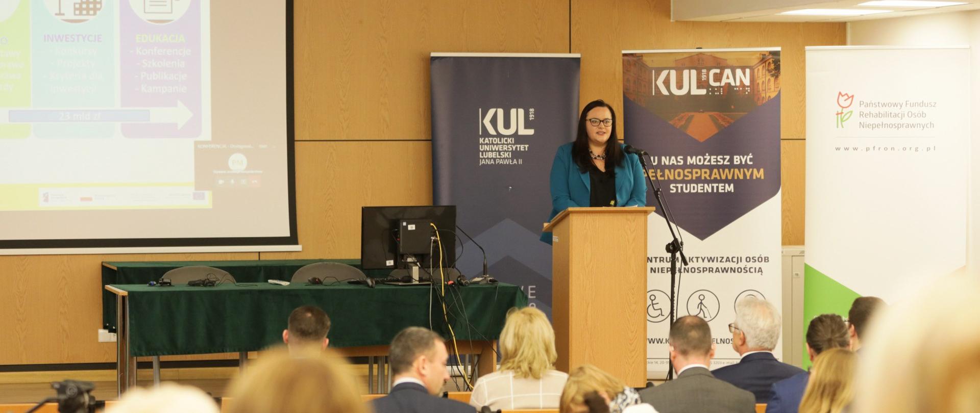 Wiceminister Małgorzata Jarosińska-Jedynak w mównicy. Przed nią publiczność. Za nią banery.