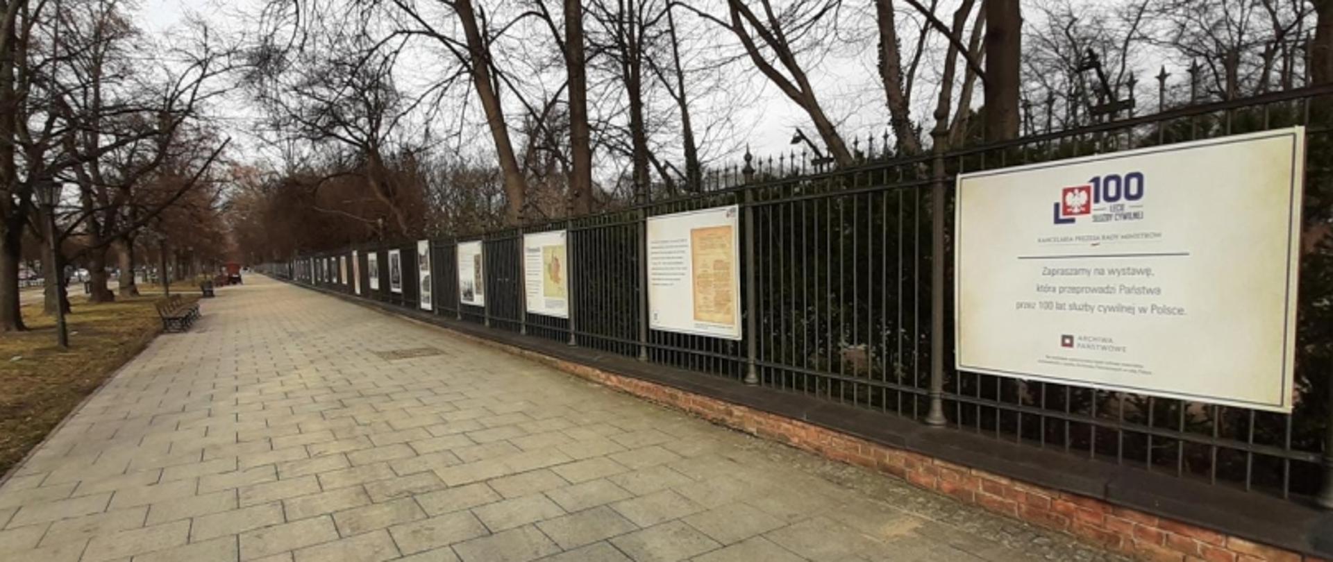 Długi chodnik obok ogrodzenia z planszami informacyjnymi, w tym plakatem z napisem „100 lat Służby Cywilnej w Polsce”; po lewej stronie ławki i drzewa bez liści.