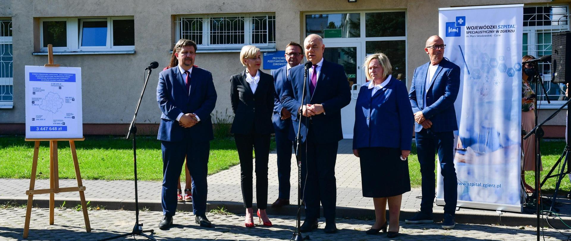 Wicepremier Jacek Sasin przemawia do mikrofonu podczas konferencji prasowej. W tle szpital w Zgierzu.
