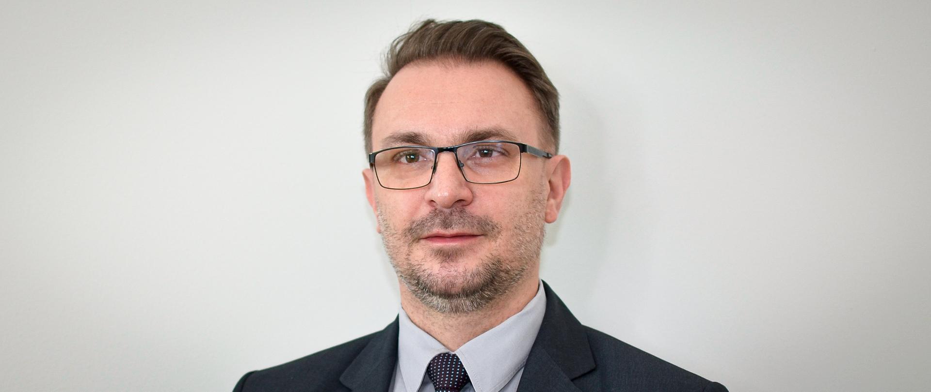 Mężczyzna w ciemnej marynarce, szarej koszuli i krawacie w biały wzorek, w okularach, z lekkim zarostem i krótkimi włosami.