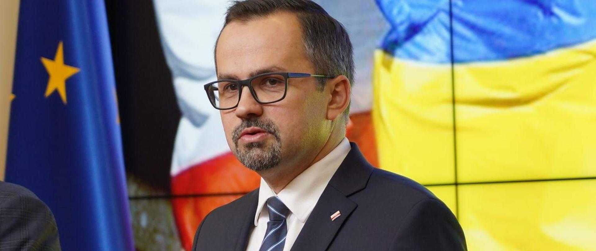 Wiceminister Marcin Horała stoi przed mikrofonem. Za jego plecami są flagi Unii Europejskiej, Polski i Ukrainy.