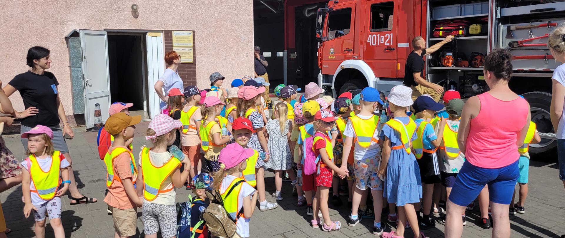 Na zdjęciu widoczny jest samochód gaśniczy koloru czerwonego wraz z dziećmi ubranymi w różnokolorowe ubrania i strażaka który jest ubrany w czarną koszulkę strażacka i opowiada o sprzęcie znajdującym się.