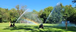 Strażacy w trakcie ćwiczenia bojowego polewają wodą tarcze, tworząc widowiskowy efekt tęczy na tle bramki piłkarskiej.