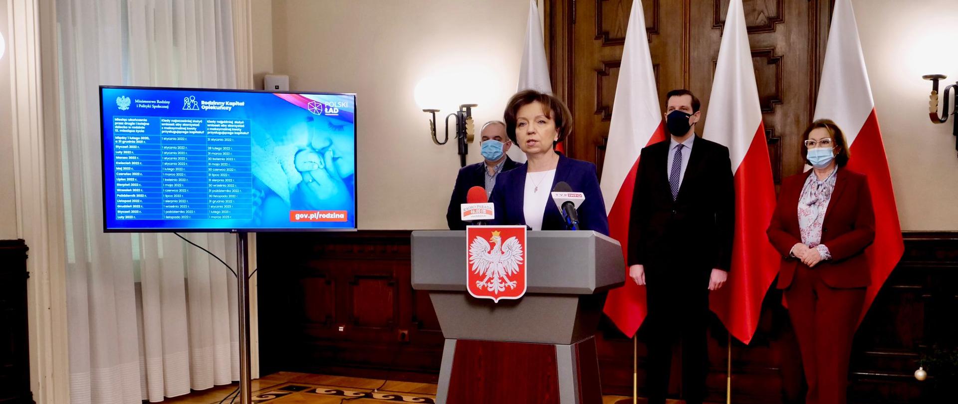 W centralnym punkcie przy mównicy z godłem Polski przemawia minister Maląg. W tle stoją trzy osoby. Za ich plecami widoczne trzy stojące biało-czerwone flagi.