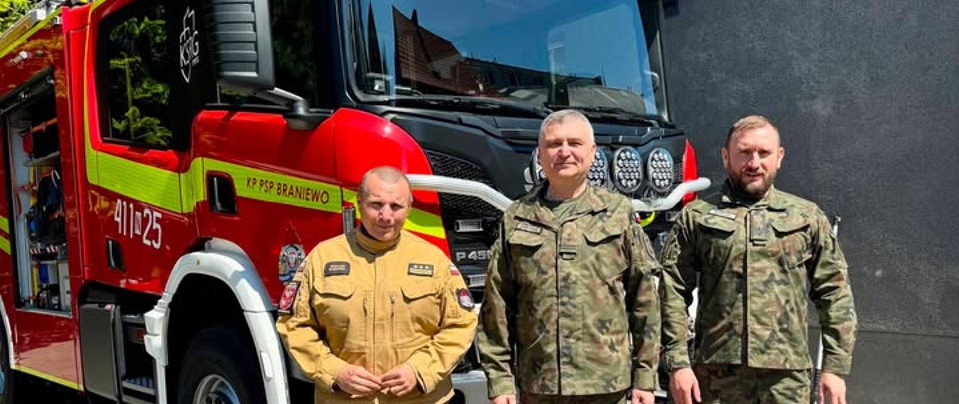 Na zdjęciu trzech mundurowych, dwóch żołnierzy i strażak w tle samochód strażacki.