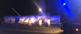 Strażacy zabezpieczają teren gdzie spadły części turbiny wiatrowej. Widać na zdjęciu bardzo duży element śmigła turbiny. Po prawej stronie wóz strażacki z włączonymi sygnałami świetlnymi w kolorze niebieskim.