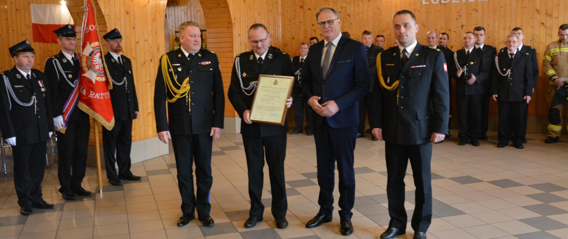 nadbrygadier Grzegorz Janowski , druh osp, mężczyzna w garniturze i strażak psp pozują do zdjęcia, po lewej poczet sztandarowy osp w tle druhowie osp