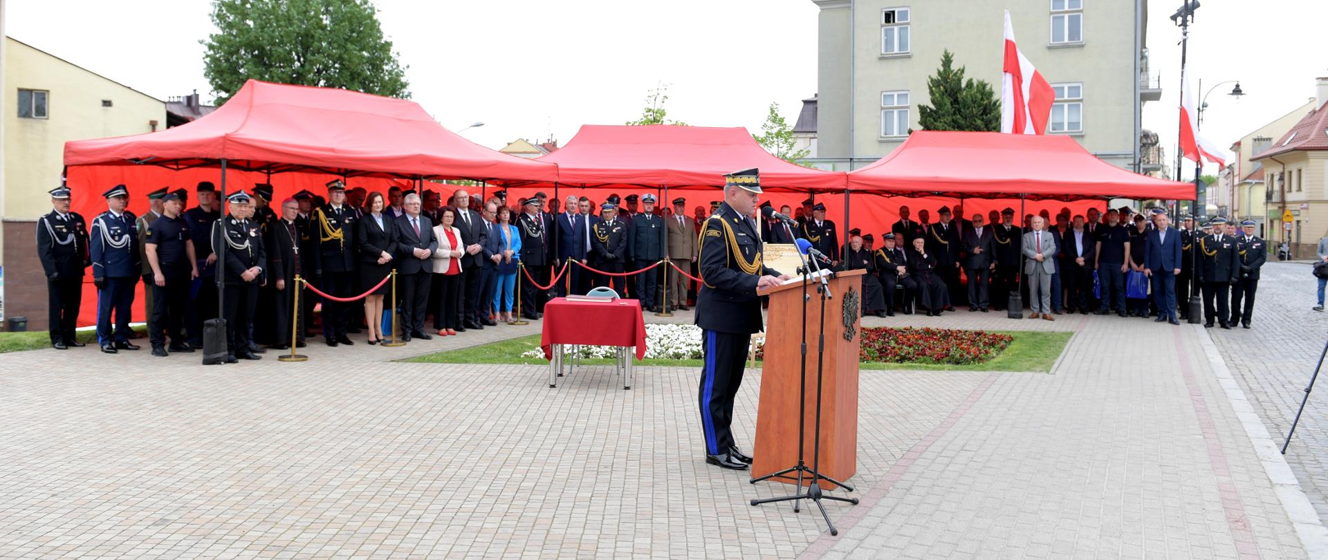 Zdjęcie przedstawia Podkarpackiego Komendanta Wojewódzkiego Państwowej Straży Pożarnej nadbrygadiera Andrzeja Babca przemawiającego przy ambonie na tle trybuny z gośćmi honorowymi.