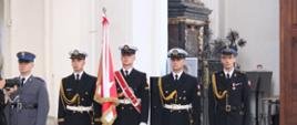 Funkcjonariusze służb mundurowych stoją obok siebie jeden z nich z szarfą przeplataną trzyma sztandar.