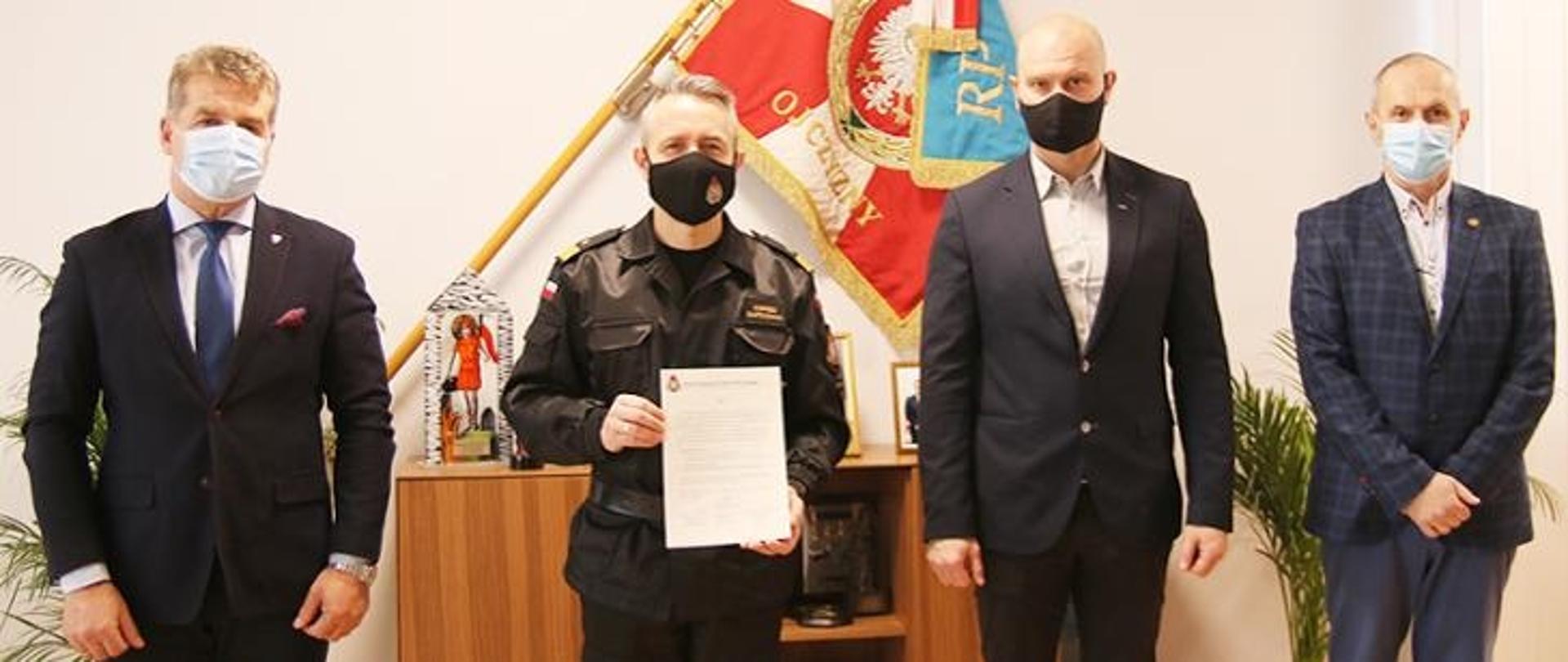 Zdjęcie przedstawia 4 mężczyzn z maseczkami na twarzy. Stoją wyprostowani. Mężczyzna drugi od prawej to Komendant Główny Państwowej Straży Pożarnej i trzyma kartkę papieru na której jest jego apel o oddawanie osocza.