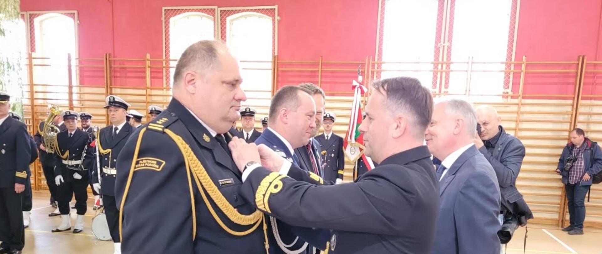 Komendant Morskiego Oddziału Straży Granicznej przypina odznaczenie zastępcy pomorskiego komendanta wojewódzkiego Państwowej Straży Pożarnej. Obok stoi funkcjonariusz Policji oraz osoba zaproszona. Obok komendanta Morskiego Oddziału Straży Granicznej stoi wojewoda pomorski.