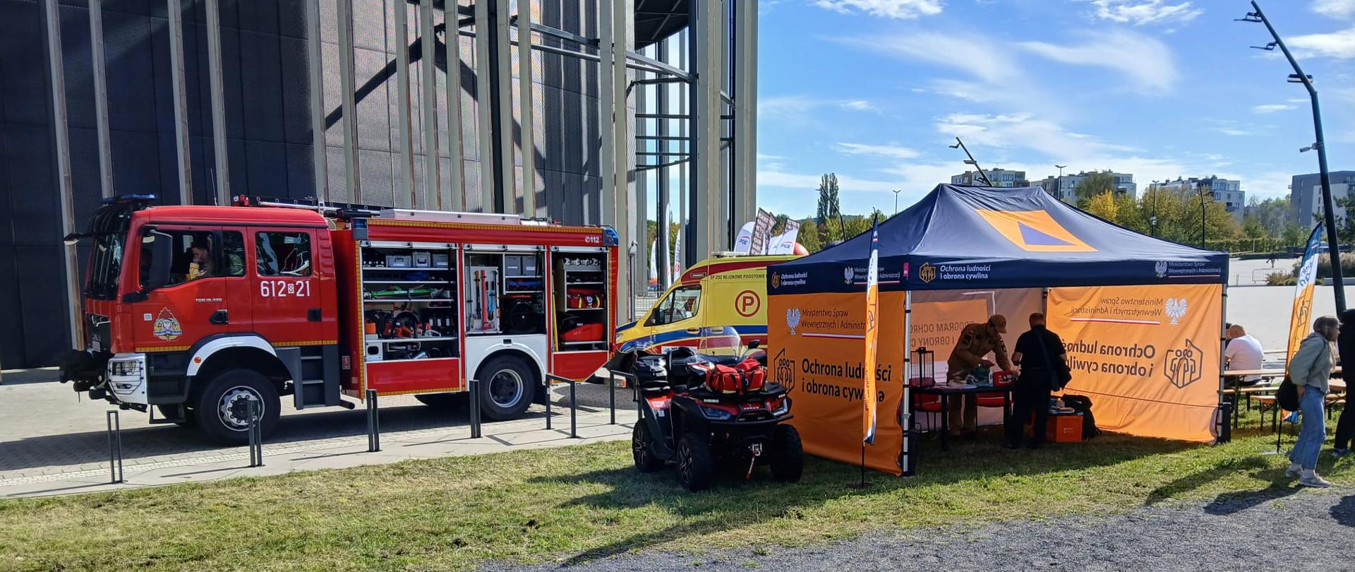 Na zdjęciu, przed dużym nowoczesnym budynkiem stadionu, znajduje się czerwony wóz strażacki oraz żółty ambulans. Obok ustawiono pomarańczowy namiot z oznaczeniami ochrony ludności i obrony cywilnej. Przed namiotem stoi quad ratowniczy, a w tle widać ludzi, flagi oraz fragment zabudowy miejskiej. Pogodny dzień, błękitne niebo.