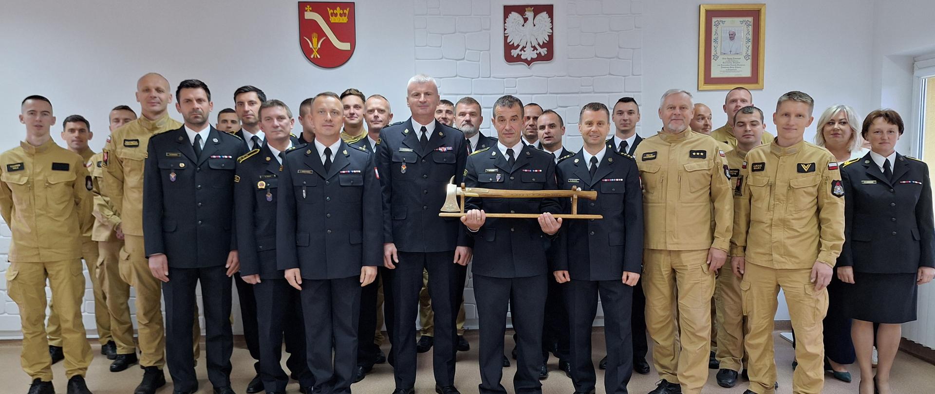 Strażacy stoją w dwuszeregu. W tle godło