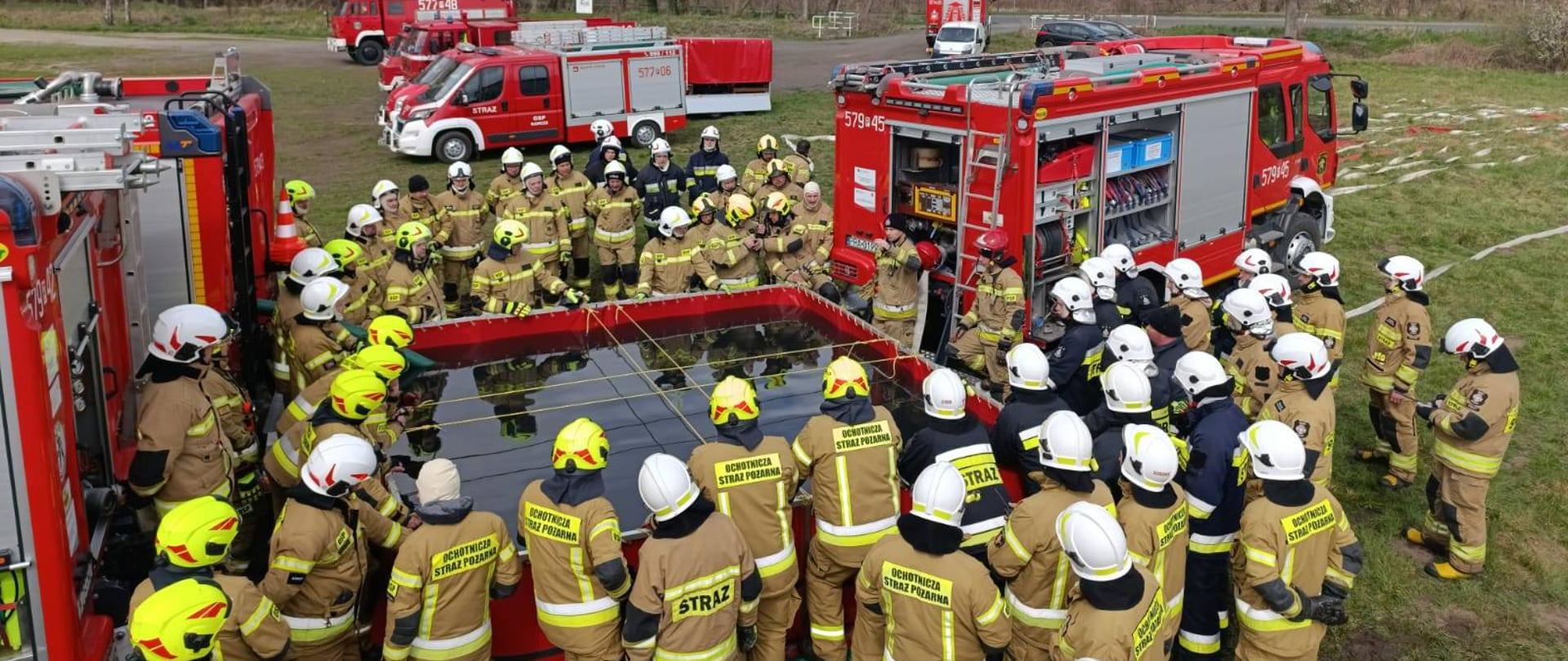 Teren zielony. Na nim, na tle drzew znajduje się kilka wozów strażackich, a także zbiornik brezentowy na wodę. Wokół zbiornika, pomiędzy samochodami stoją strażacy uczestniczący w warsztatach. Maja na sobie ubrania specjalne w kolorach piaskowym oraz czarnym, a także białe i żółte hełmy. Z prawej strony widać rozwinięte węże.