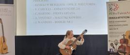 Na zdjęciu uczennica grająca na gitarze podczas konkursu - II Ogólnopolski Konkurs Gitarowy o tytuł "Mistrza Sześciu Strun" - 11 luty 2023, w tle widoczny program, a po prawej stronie baner konkursu