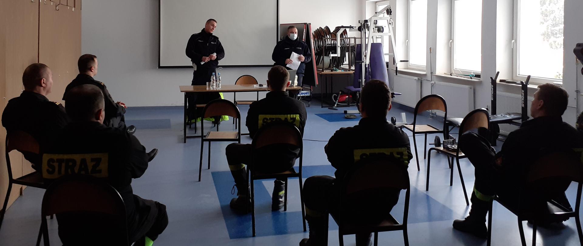 Na pierwszym planie widać siedzących tyłem na krzesłach strażaków. W dalszej części widać dwóch stojących policjantów, przed którymi ustawiony jest stolik, a na nim komputer. Wszyscy znajdują się w pomieszczeniu budynku.