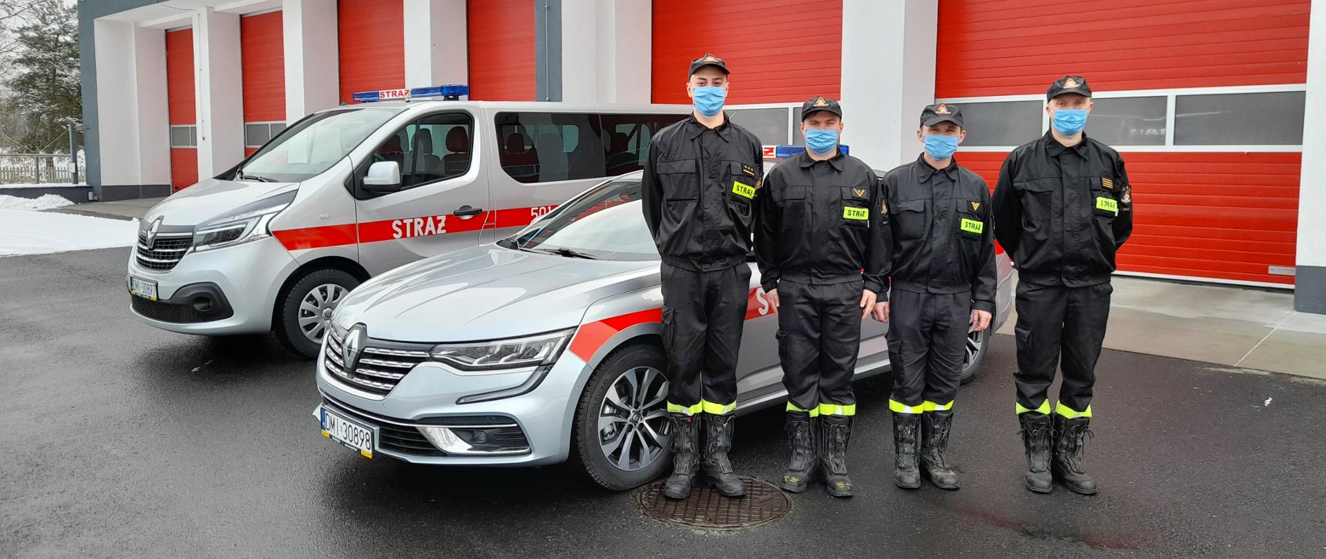 Czterech strażaków w ubraniach koszarowych stoi na tle samochodów osobowych i garaży jednostki ratowniczo gaśniczej w Miliczu