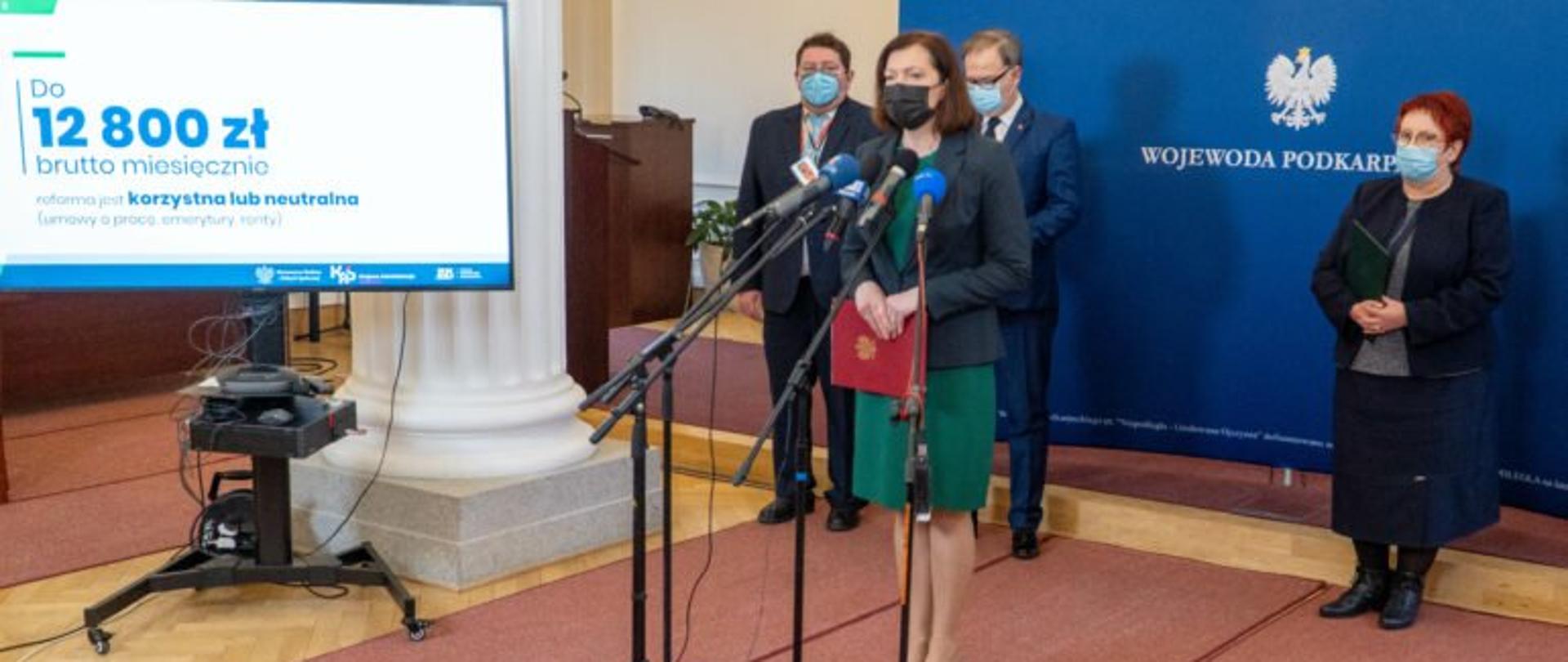 Wojewoda podkarpacki Ewa Leniart zabiera głos podczas konferencji prasowej. Za nią stoją od lewej zastępca dyrektora Izby Administracji Skarbowej w Rzeszowie Jacek Cichy, dyrektor Izby Administracji Skarbowej w Rzeszowie Witold Mrozek i dyrektor ZUS oddział w Rzeszowie Ewa Papiewska-Borkowska. Po lewej stronie wojewody fragment prezentacji na ekranie.