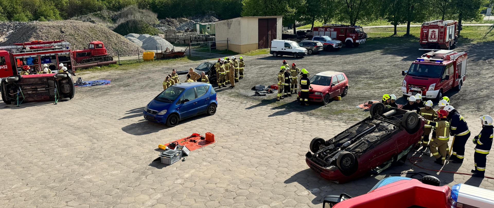 Druhów stojących przy wrakach pojazdów. Przy każdym stanowisku instruktor tłumaczy zasady wykonywania dostępu. Samochody są ustabilizowane. Na placu stoją także wozy strażackie.