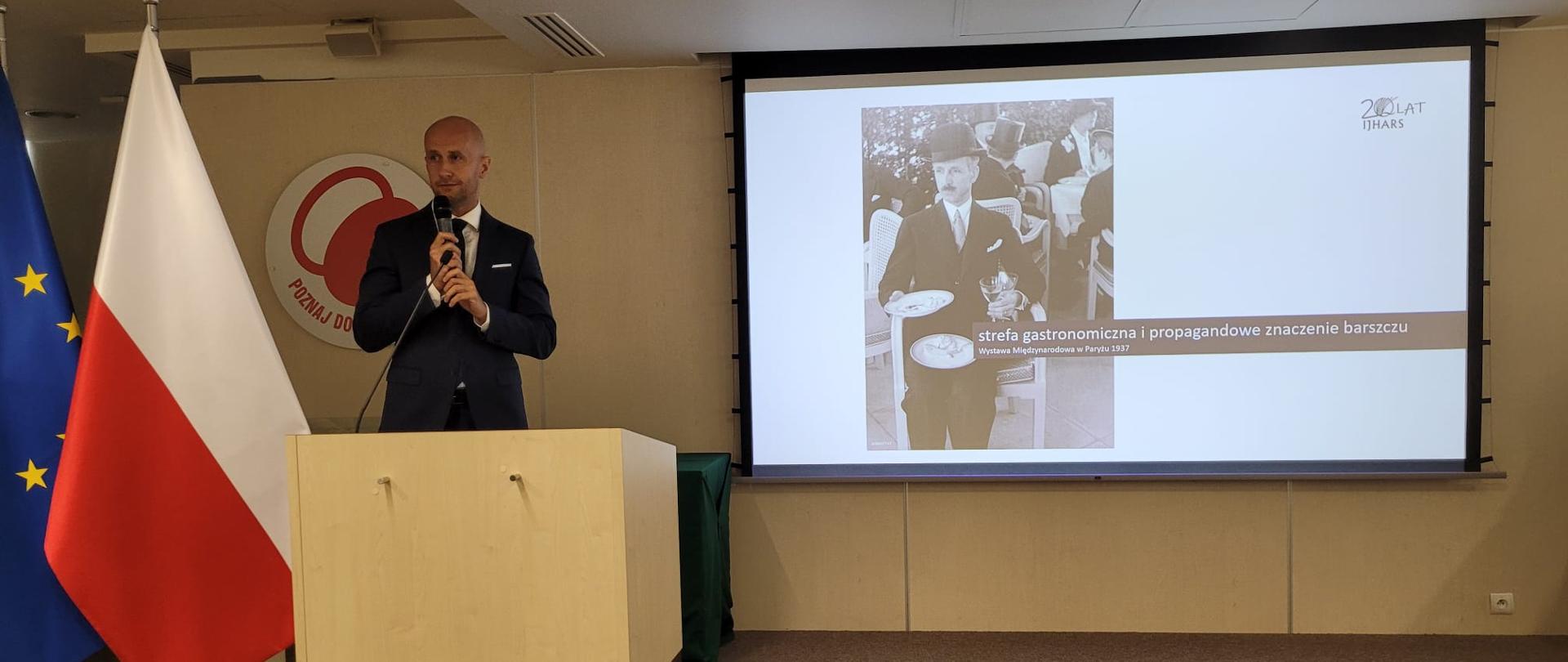 Na mównicy z mikrofonem w dłoni stoi Tomasz Michalski. Po jego prawej stronie flaga polska i Unii Europejskiej. Po prawej ekran ze slajdem przedstawiającym mężczyznę w meloniku niosącym 2 pełne talerze i 2 napełnione kieliszki do wina. Napis: strefa gastronomiczna i propagandowa znaczenie barszczu. Wystawa Międzynarodowa w Paryżu 1937