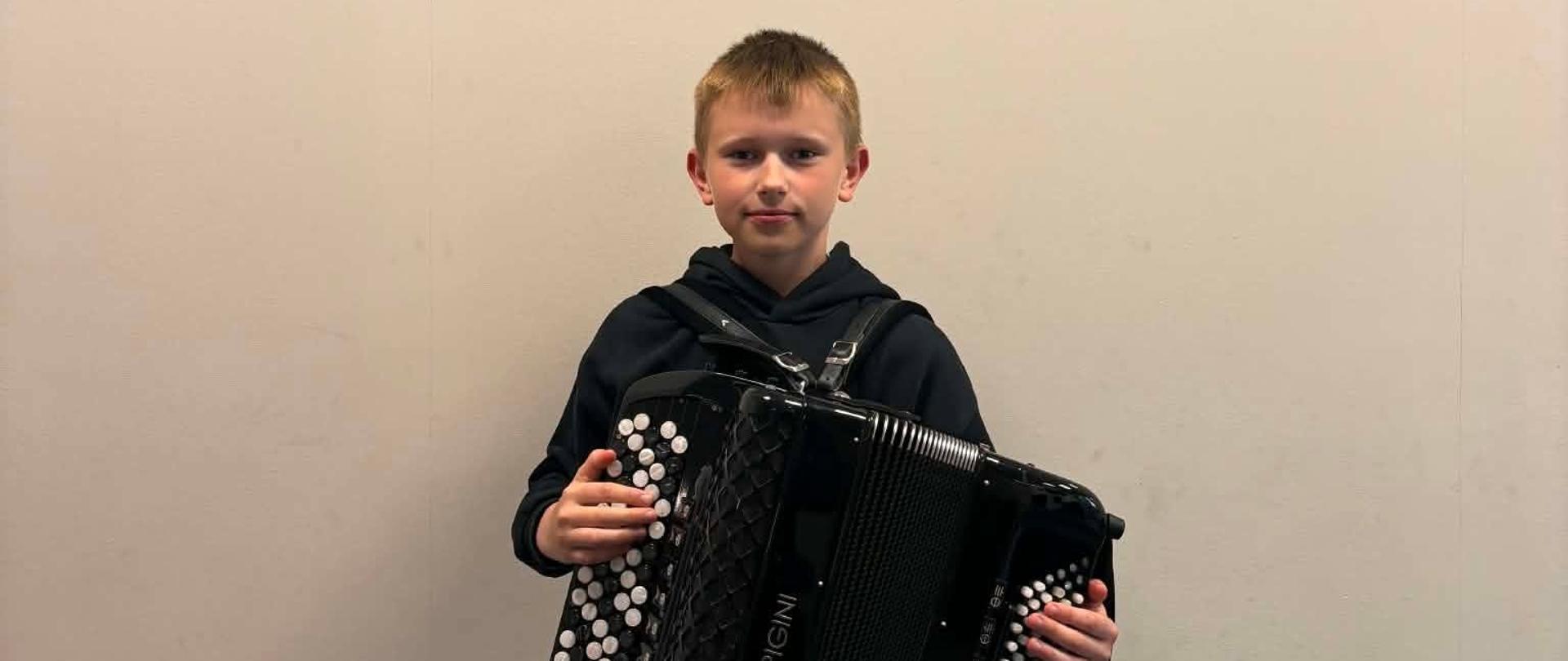 Na zdjęciu widać chłopca, który trzyma akordeon. Na ścianie wisi panel akustyczny z grafiką instrumentu. 