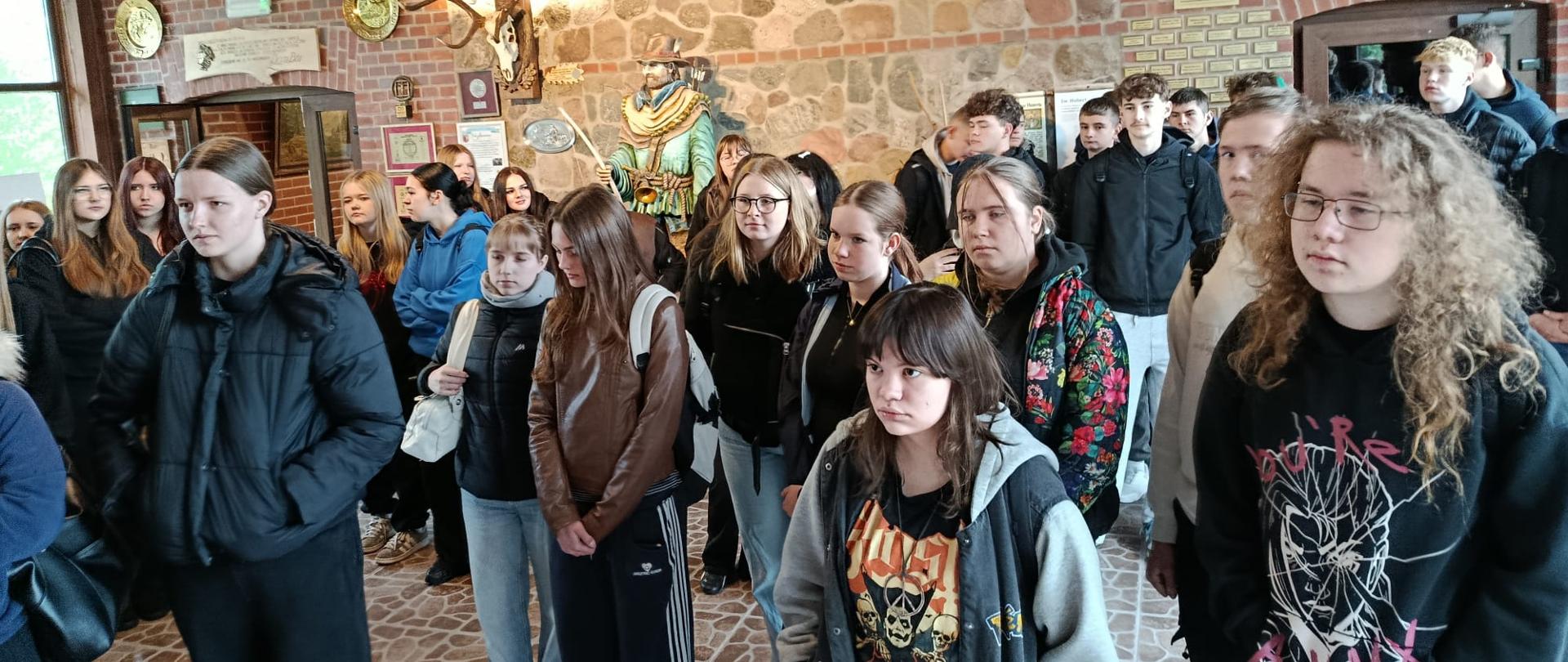 Duża grupa uczniów ZSCKR w Samostrzelu stoi w sali muzealnej o kamienno-ceglanych ścianach, słuchając przewodnika. W tle widoczne są dekoracje myśliwskie, poroża oraz rzeźba przedstawiająca postać w tradycyjnym stroju.