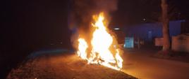 Zdjęcie przedstawia objęty pożarem samochód osobowy w porze nocnej. Samochód zaparkowany jest na terenie zielonym.