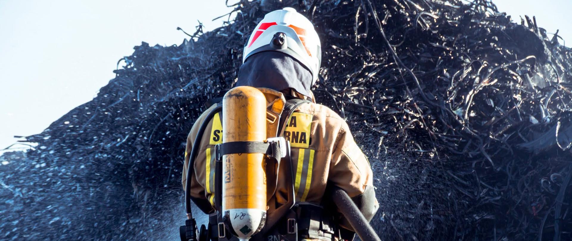 Na pierwszym palnie strażak odwrócony plecami w umundurowaniu bojowym, z aparatem ODO, gasi wielką hałdę czarnego materiału, brak ognia, dzień