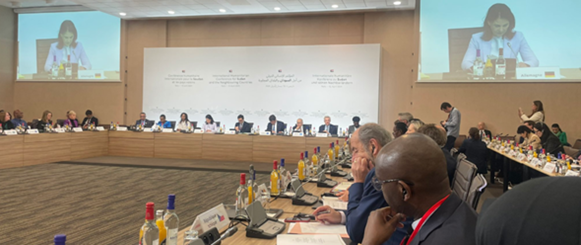 Delegaci w trakcie konferencji ze stołem prezydialnym i dwoma dużymi ekranami na których widać przemawiającą kobietę.