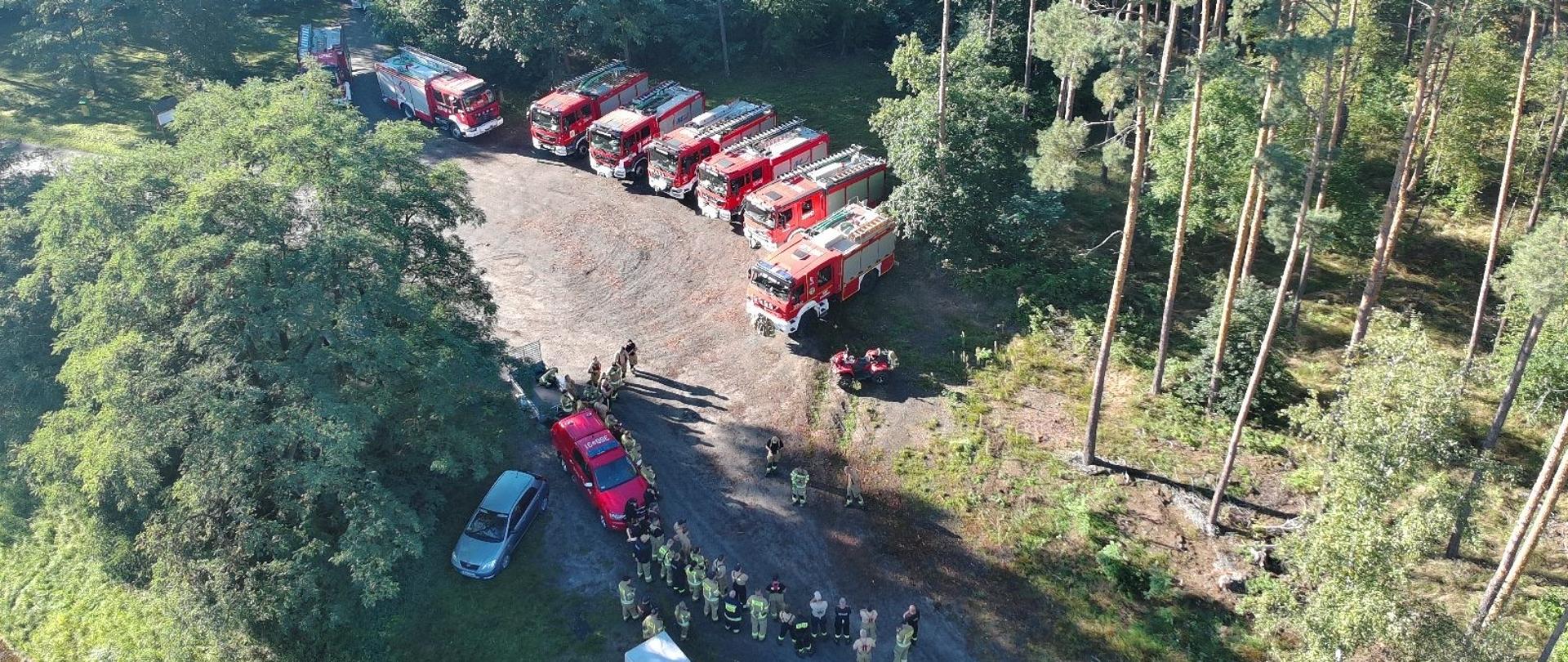 Zdjęcie przedstawia samochody pożarnicze oraz zbiórkę strażaków przed rozpoczęciem ćwiczeń. Pojazdy ustawione są w szeregu, a zdjęcie wykonane jest z drona.
