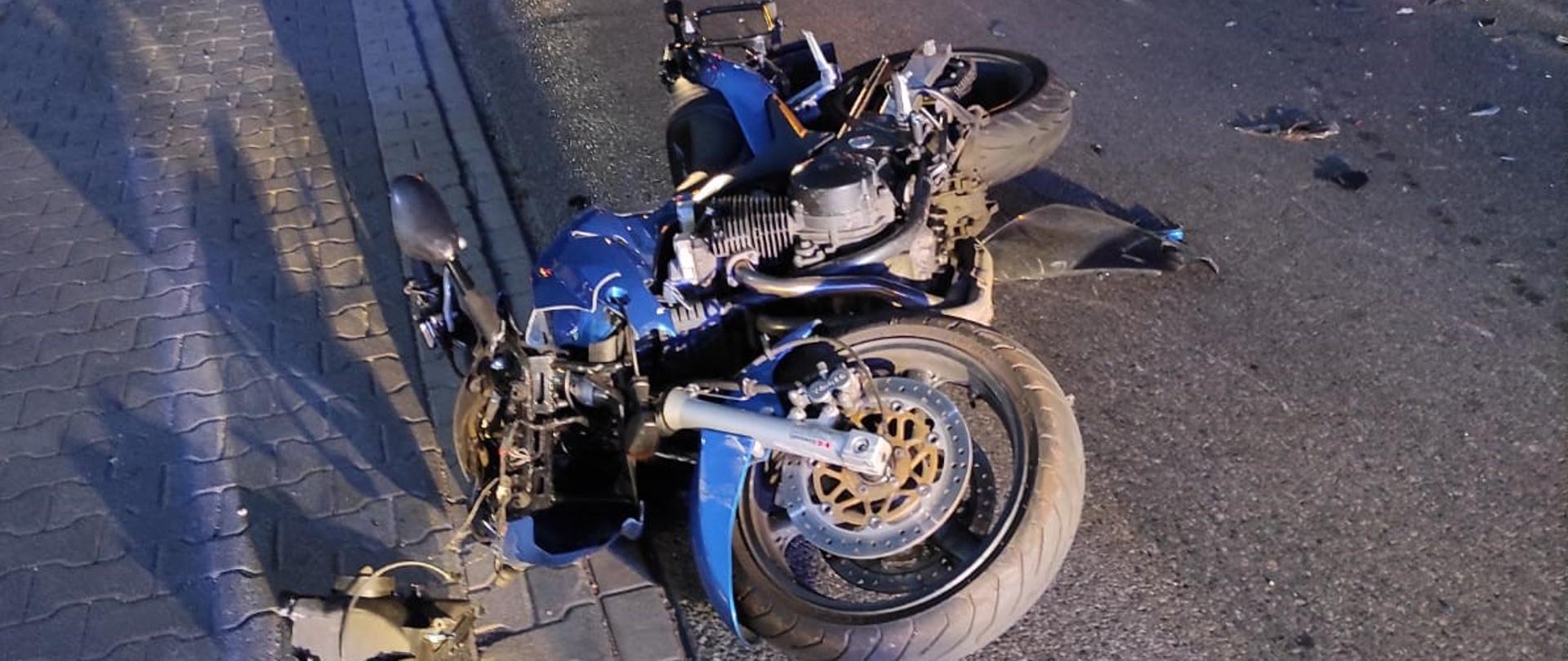 Zdjęcie wykonano w godzinach późno wieczornych. Na pasie ruchu leży rozbity motocykl po zderzeniu z samochodem. Na drugim planie zabudowania miejscowości i pojazdy przemieszczające się drogą powiatową.