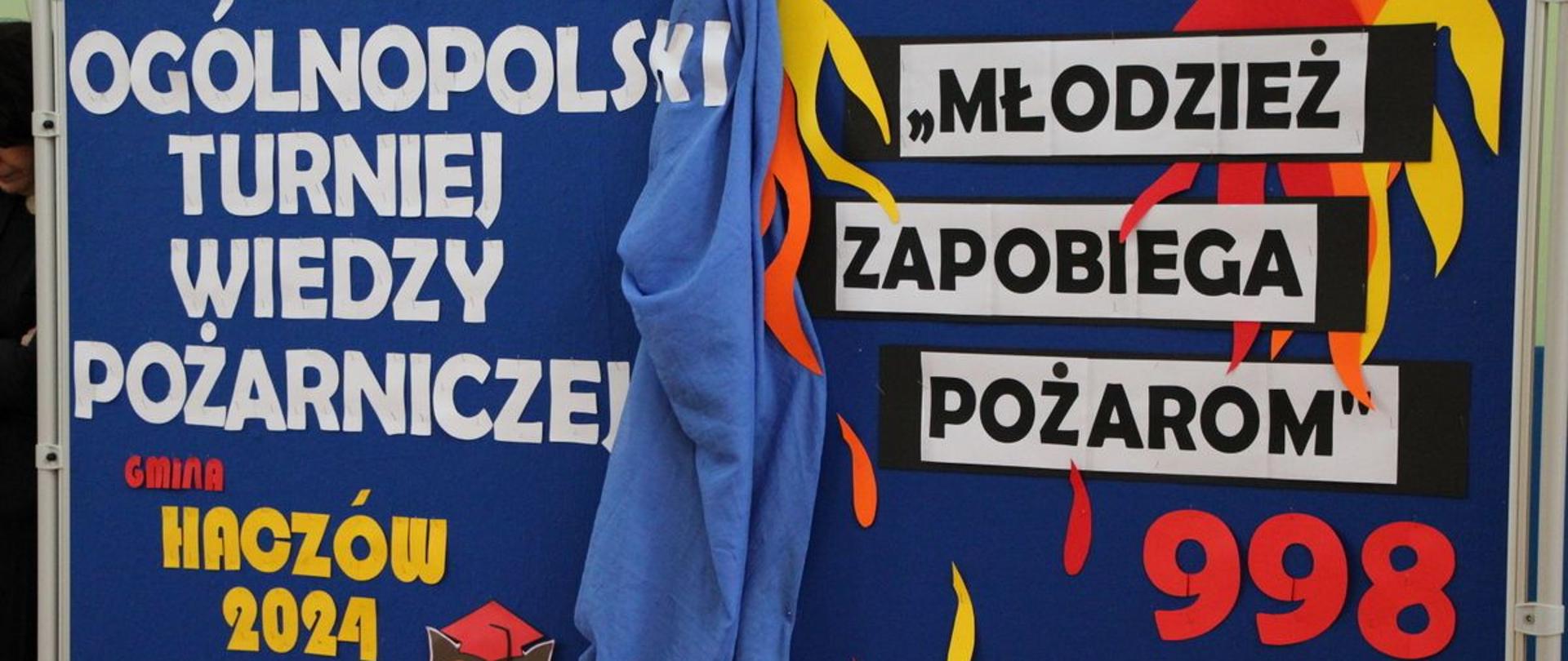 Kolorowa fotografia przedstawiająca baner z napisem „Ogólnopolski Turniej wiedzy pożarniczej” Gminy Haczów 2024, „Młodzież zapobiega pożarom z dopisem 998”