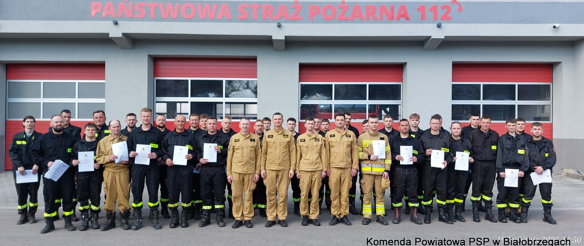 Zdjęcie grupowe uczestników szkolenia podstawowego OSP oraz instruktorów przed budynkiem Komendy Powiatowej PSP w Białobrzegach. Strażacy w mundurach trzymają otrzymane zaświadczenia.