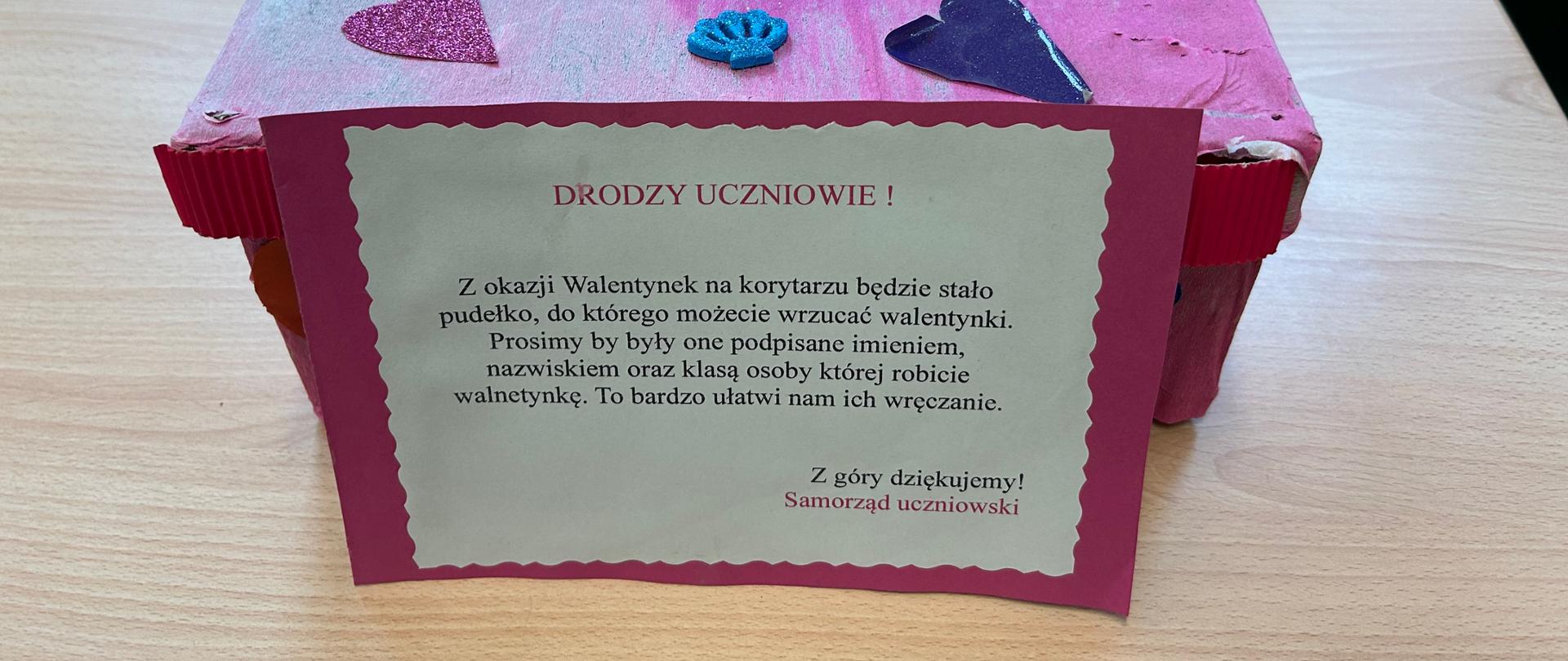 Zdjęcie przedstawia pudełko, do którego można wrzucać walentynki. Pudełko stoi na stole. Ozdobione jest różową bibułą oraz serduszkami. Na górze wycięty jest otwór, ozdobiony wokół cekinami. Przed pudełkiem znajduje się kartka z informacją od samorządu uczniowskiego o treści: " Drodzy Uczniowie! Z okazji Walentynek na korytarzu będzie stało pudełko, do którego możecie wrzucać walentynki. Prosimy by były one podpisane imieniem i nazwiskiem oraz klasą osoby której robicie walentynkę. To bardzo ułatwi nam ich wręczanie. Z góry dziekujemy! Samorząd uczniowski."