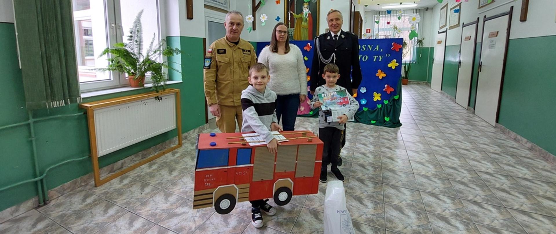 Kolorowa fotografia wykonana w pomieszczeniu przedstawiająca umundurowanych strażaków wraz z dziećmi, które otrzymały nagrody w konkursie plastycznym.