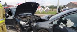 Zdjęcie przedstawia dwa samochody osobowe po zderzeniu czołowym. W samochodzie po prawej stronie widać poduszkę powietrzną po wystrzeleniu. Po lewej stronie stoi strażak.