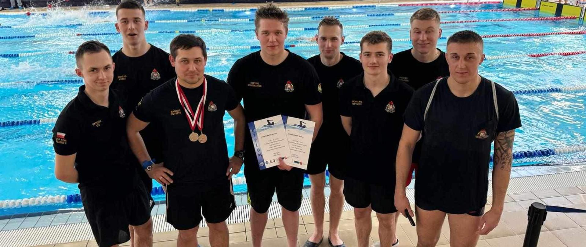 Pamiątkowe zdjęcie grupowe (8 osób) trenera oraz kadetów Centralnej Szkoły PSP uczestniczących w zawodach pływackich, w tle pływalnia