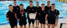 Pamiątkowe zdjęcie grupowe (8 osób) trenera oraz kadetów Centralnej Szkoły PSP uczestniczących w zawodach pływackich, w tle pływalnia