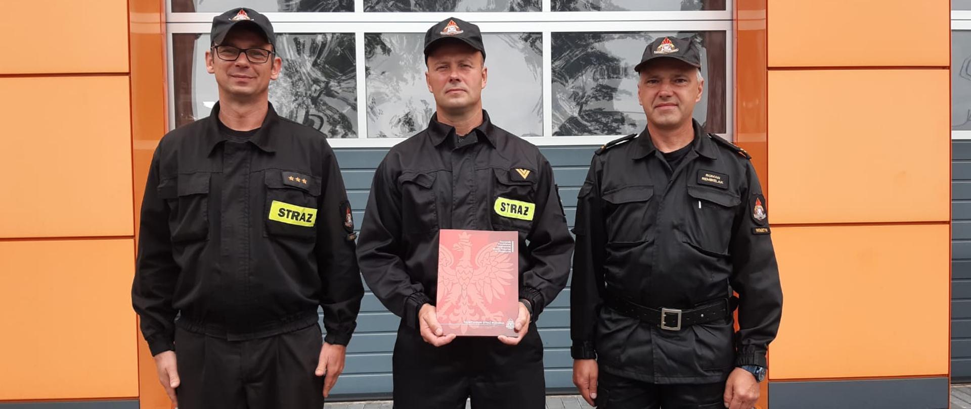 Na zdjęciu funkcjonariusz trzymający teczkę. Obok niego stoi Komendant Powiatowy PSP w Wolsztynie oraz dowódca Zmiany. W tle garaż JRG. Zdjęcie zrobione na zewnątrz, na placu komendy.