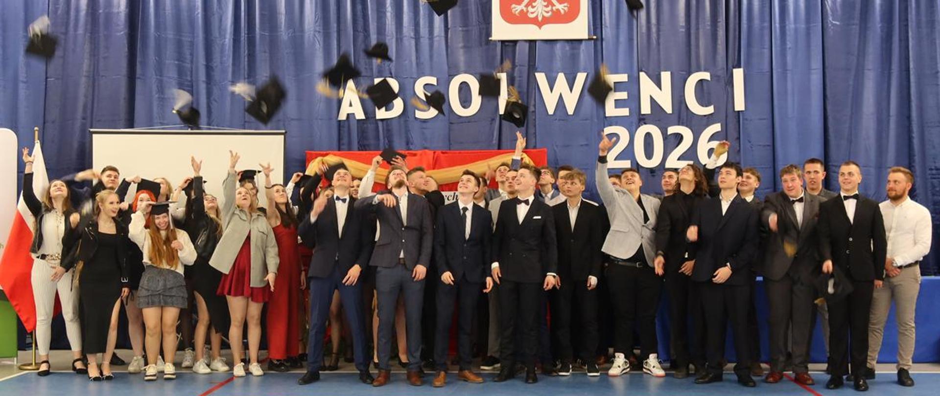 Duża grupa dziewcząt i chłopaków stojących w sali gimnastycznej w galowych strojach, niektórzy podrzucają birety. W tle napis: "Absolwenci 2026".