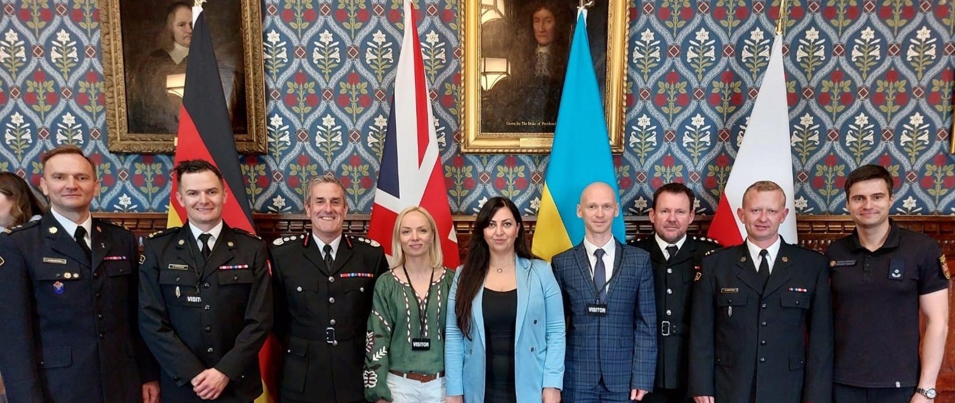 6 strażaków (po 3 z lewej i prawej strony) pomiędzy nimi 2 kobiety pozują na stojąco do zdjęcia, z animi flago od lewej Wlk Brytanii, Ukrainy i Polski, na ścianie wiszą dwa obrazy